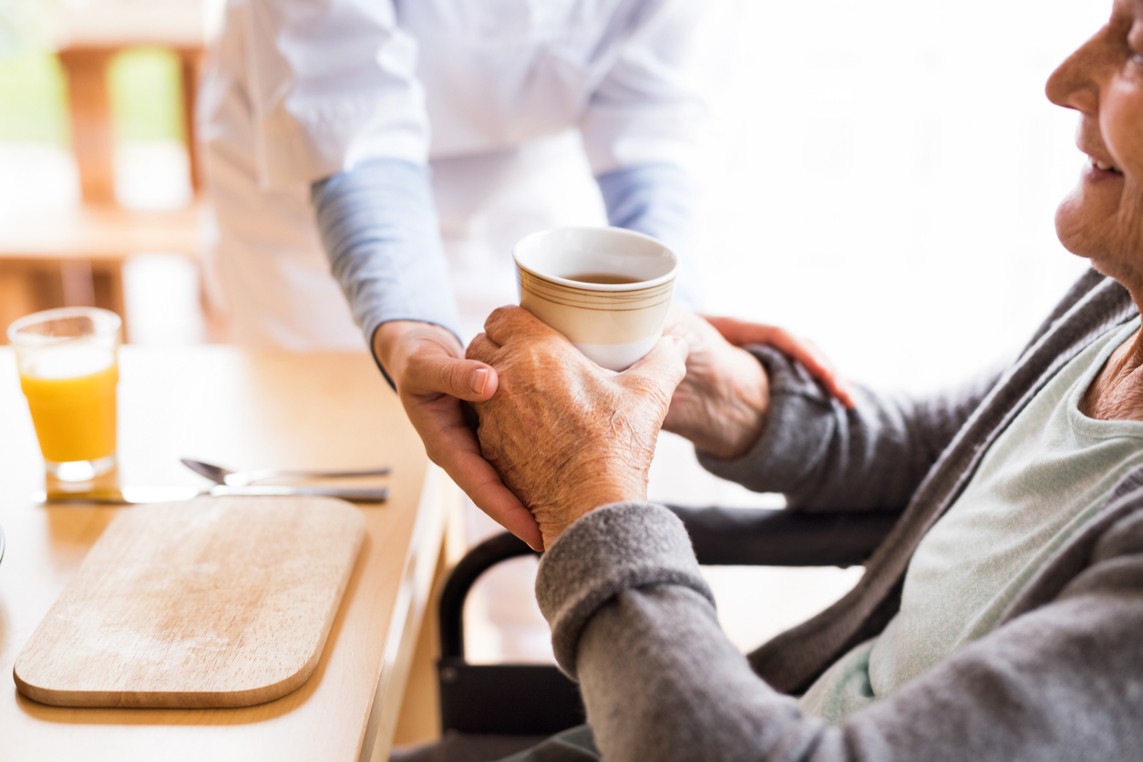 Eine Krankenschwester reicht einer älteren Frau, die am Tisch sitzt, eine Tasse.